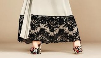 por baixo de uma túnica creme com renda preta aparecem os pés da modelo calçando sapatos de saltos altos com flores multicoloridas trabalhadas e aplicadas no corpo do calçado em diferentes texturas.