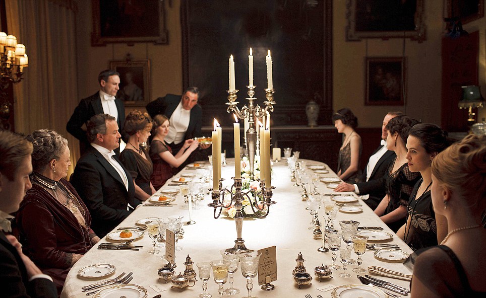 mesa clássica de uma família inglesa - do seriado Downton Abbey - os homens usando casacas pretas e as mulheres usando vestidos longos. Ao centro dois candelabros de cinco velas acesas.