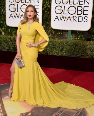 A atriz Jennifer Lopez usa vestido longo, cor amarelo ovo, com um tipo de mini capa sobreposta da mesma cor. Os seus cabelos estão clareados quase loiro, cor de batom vermelho e ela usa uma bolsa em prata.