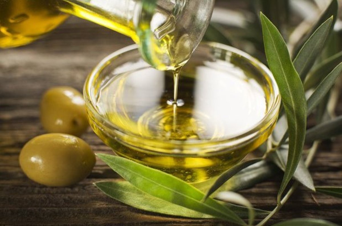 pote cheio de azeite de oliva e uma garrafa despeja mais azeite, ao lado duas azeitonas verdes e do outro lado ramos de oliveira.