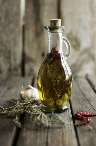 Sobre um engradado de madeira uma garrafa de vidro transparente com azeite de oliva e algumas ervas e pimenta. Ao lado da garrafa, algumas ervas , uma cabeça de alho e 3 pimentas dedo-de-moça