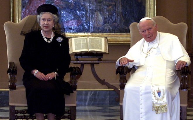 Em audiência papal no Vaticano. Rainha Elizabeth II usa vestido cor preta, está sentada a esquerda , ao centro um biblia aberta sobre uma mesa e a direita o Papa João Paulo II, sentado usando o seu traje tradicional todo branco.