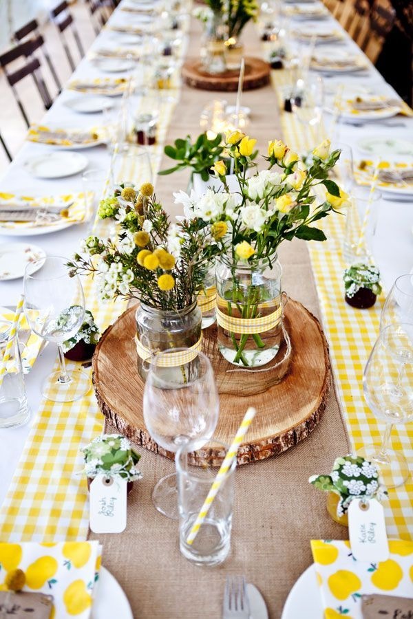 mesa longa em madeira clara com toalha branca rústica quadriculada de amarelo claro e no centro arranjos improvisados de flores do campo amarelas em vasinhos improvisados de vidro reciclado