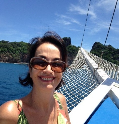 Claudia Matarazzo, usando óculos escuros , grandes, está junto a proa de um veleiro que navega pelo mar de Fernando de Noronha, num dia límpido de muito sol.