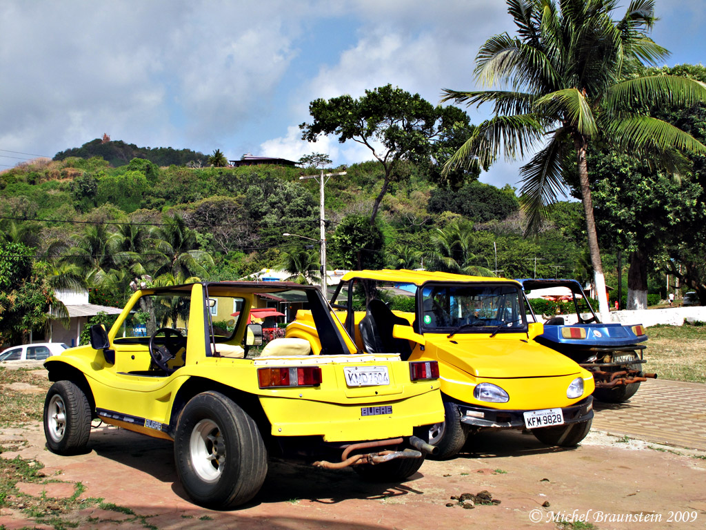 Em Fernando de Noronha, junto a uma mata nativa, tres buggys estacionados. dois amarelos e um azul. aguardam clientes para os passeios pelas praias e região.