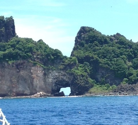 Imagem da gruta de Fernando de Noronha vista de um veleiro que navega pela região. Ao fundo montanhas com muita vegetação e entre elas uma pequena gruta. O mar no seu azul brilhante .
