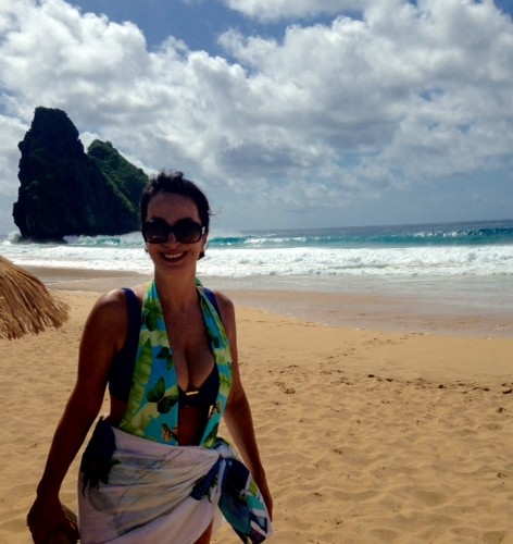Claudia Matarazzo, numa das praias de Fernando de Noronha, usa cabelos presos , usa também uma saida de praia florida e um top azul. Usa óculos escuros grande e sorri.
