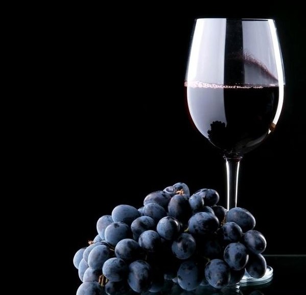 uma taça de vinho tinto bem escuro está contra um fundo negro e um cacho com uvas também escuras cor de beringela está ao pé da taça. O efeito é de impacto com o contraste dos tons escuros e do brilho do cristal.