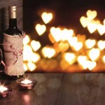 Sobre uma mesa de madeira está uma garrafa de vinho envolta em uma capa de juta rústica com das velas bem baixinhas a sua frente e taças. Ao fundo luzes em forma de coração iluminam o ambiente.