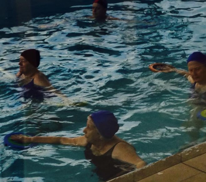Numa piscina coberta, algumas senhoras nadam, usando uniforme (maiô) na cor azul, e usam tocas da mesma cor.
