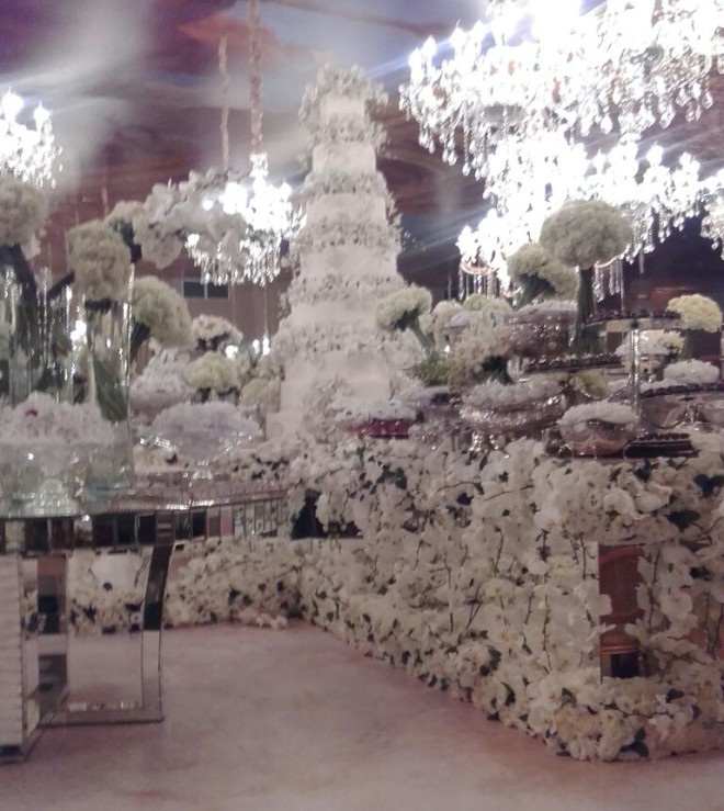 Um salão de festa mostra a mesa de doces com doces em toda a sua superfície e embaixo dela flores e mais flores brancas. Junto com os doces também há flores arranjadas de modo a se confundir com os doces brancos. Ao fundo e em uma mesa em primeiro plano mais flores praticamente ocupando todo o espaço livre do salão. Um super lustre de cristal está pendurado acima da mesa quase tocando nas flores.