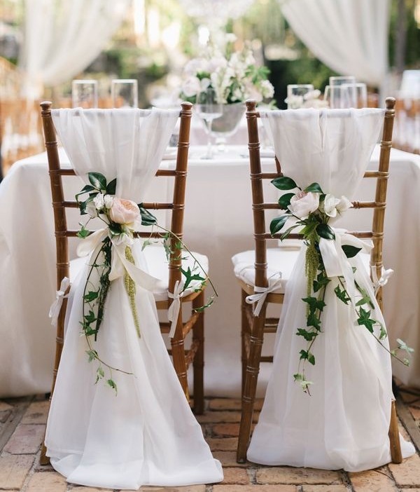 duas cadeiras em frente a uma mesa de jantar de noivos estão vestidas com um pesado tecido branco que cai drapeado pelo encosto arrastando no chão e arrematado por um pequeno buquê de rosas brancas.