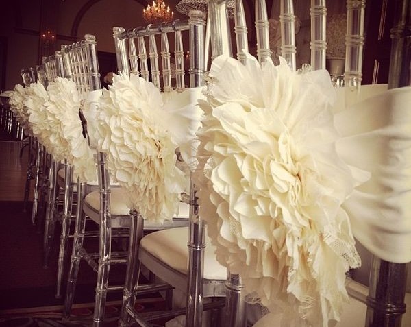Em uma decoração de casamento cadeiras de acrílico estão enfileiradas em frente a uma mesa longa ecom flores em tecido branco gigantes, que cobrem praticamente todo o encosto amarradas no mesmo.