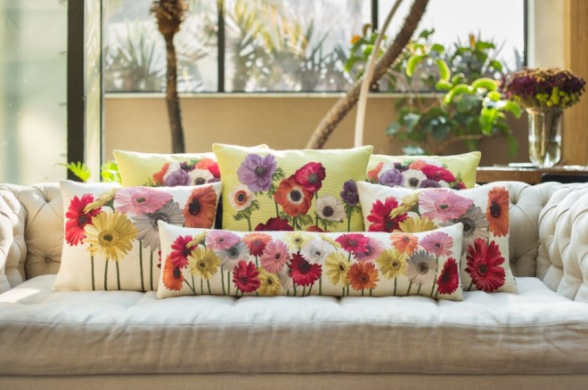 Sobre um sofá de cor creme colocado em frente a uma janela , estão colocadas várias almofadas de fundo também creme porém estampadas com enormes flores de todas as cores. O efeito é de alegria e muita cor.