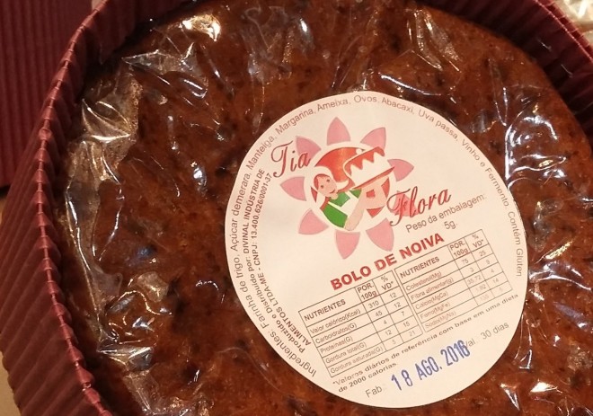 A foto mostra bem de perto um bolo redondo de cor marrom chocolate embrulhado em papel celofane protegido por uma apara de papelão sanfonado ao redor. É uma embalagem comercial pronta pra ser vendida.