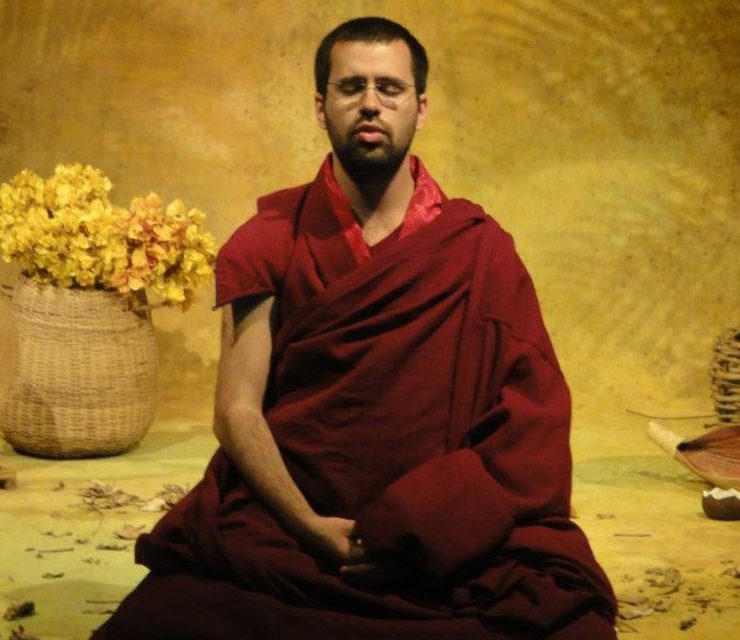Lama Michel Rimpoche, monge budista, jovem, magro, cabelos raspados, sentado na posição de seiza, medita num templo florido, usando os mantos marsala, simbolos desta religião. Está de olhos fechados.