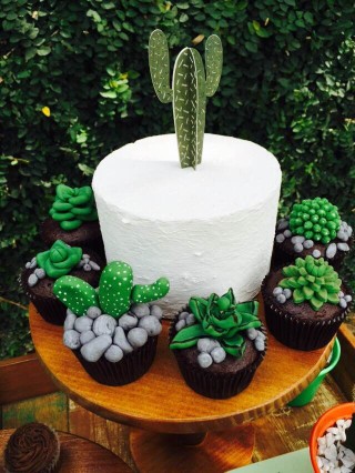 Uma mesa pequena e redonda apoia um prato de bolo branco com um cactus de açúcar verde confeitado enfeitando. Ao redor pequenos vasos de cactos naturais compõem o visual.