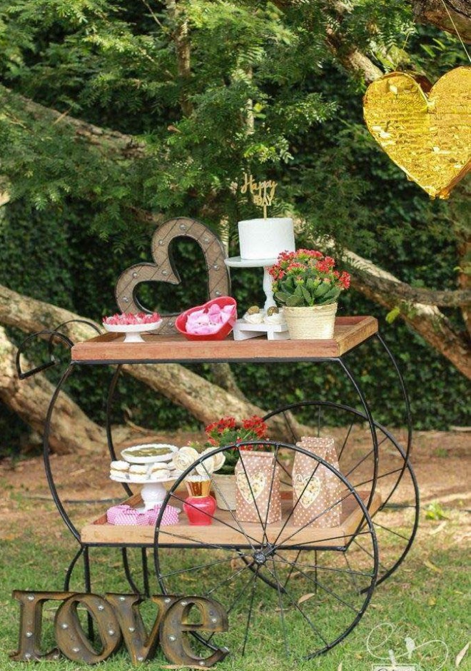 Um carrinho de chá está colocado em um ponto do jardim com doces e bolso em sua superfície superior e também na prateleira de baixo.. O efeito é delicado e rústico.