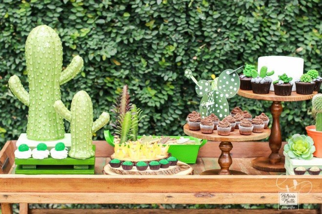 Um aparador de madeira médio está com várias travessas de doces e uma boleira todos de tamanho pequeno ou médio. Alguns tem formato de cactos de açúcar, outros de docinhos e o há também cupcakes - ou mini bolos