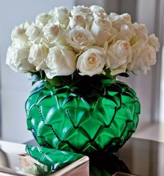 Um vaso de flores de vidro verde folha trabalhado em losangos baixo e gordinho está cheio de rosas brancas e se reflete em uma caixa de tampa de espelho ao seu lado. O efeito da decoração é elegante e alegre