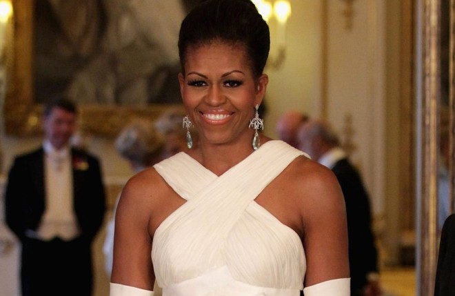 Michelle Obama veste um modelo de vestido branco sem ombro mas com alças largas cruzadas na frente. Sua pele negra contrasta com o tom creme claro do tecido e ele está de cabelo preso em um coque clássico.