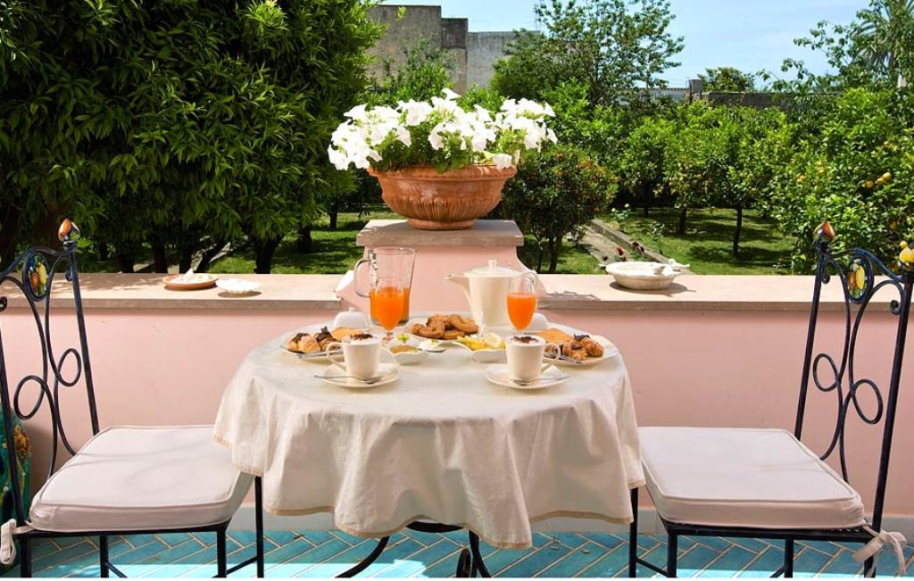 mesa de café da manhã, numa sacada de um hotel em Capri, Italia, num belíssimo cenário da italiano. Sobre a mesa com toalha com tecido claro, temos copos de sucos, xícaras , pães e ao fundo , flores num vazo .