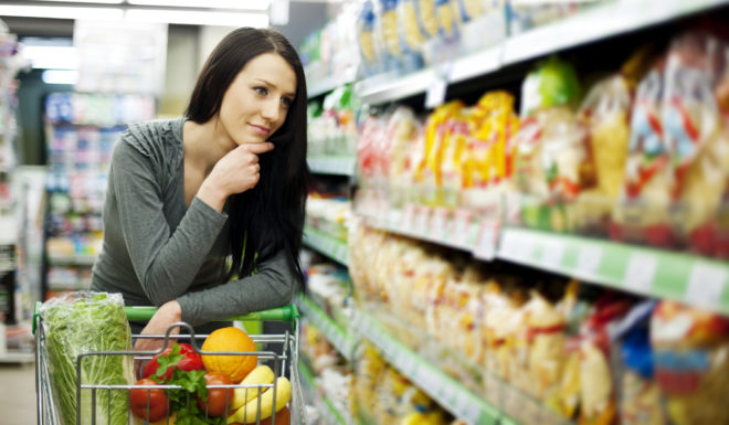 Num supermercado, mulher de cabelos longos, cor castanho escura, debruça sobre o carrinho de compras, e olha as gôndolas num supermercado.