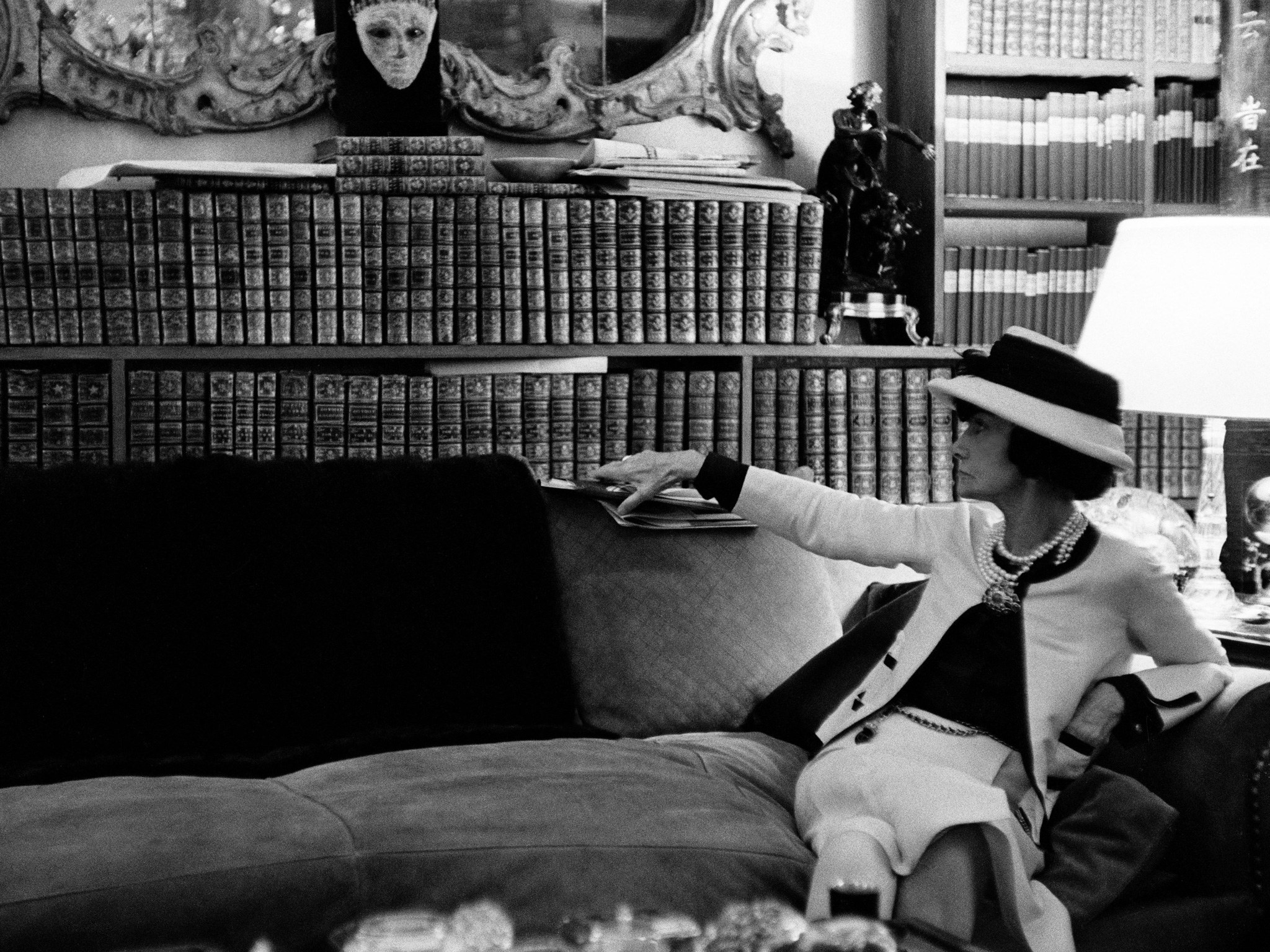 A estilista Coco Chanel - na sala de sua casa-atelier- sentada no sofá - Ela usa chapéu e um conjunto claro. Ao fundo uma prateleira com diversos livros