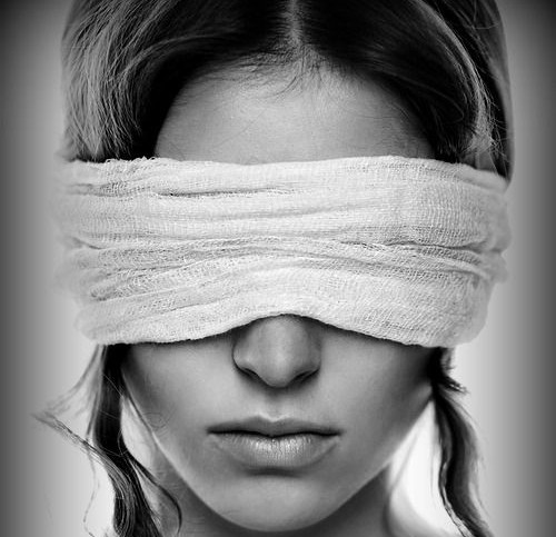 Rosto de um mulher com os olhos vendados por um pano branco. Ela tem tranças de seus cabelos nas laterais da cabeça.