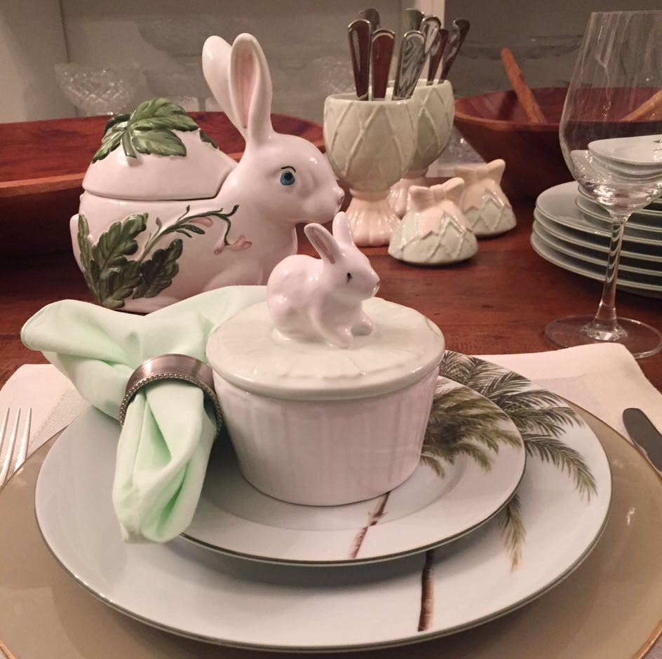 sobre um prato branco com palmeiras verdes está uma tijelinha de sopa com tampa de coelhinho no puxador. Atrás vemos um outro coelho maior em porcelana branca e ao fundo ovos e outros pratos.