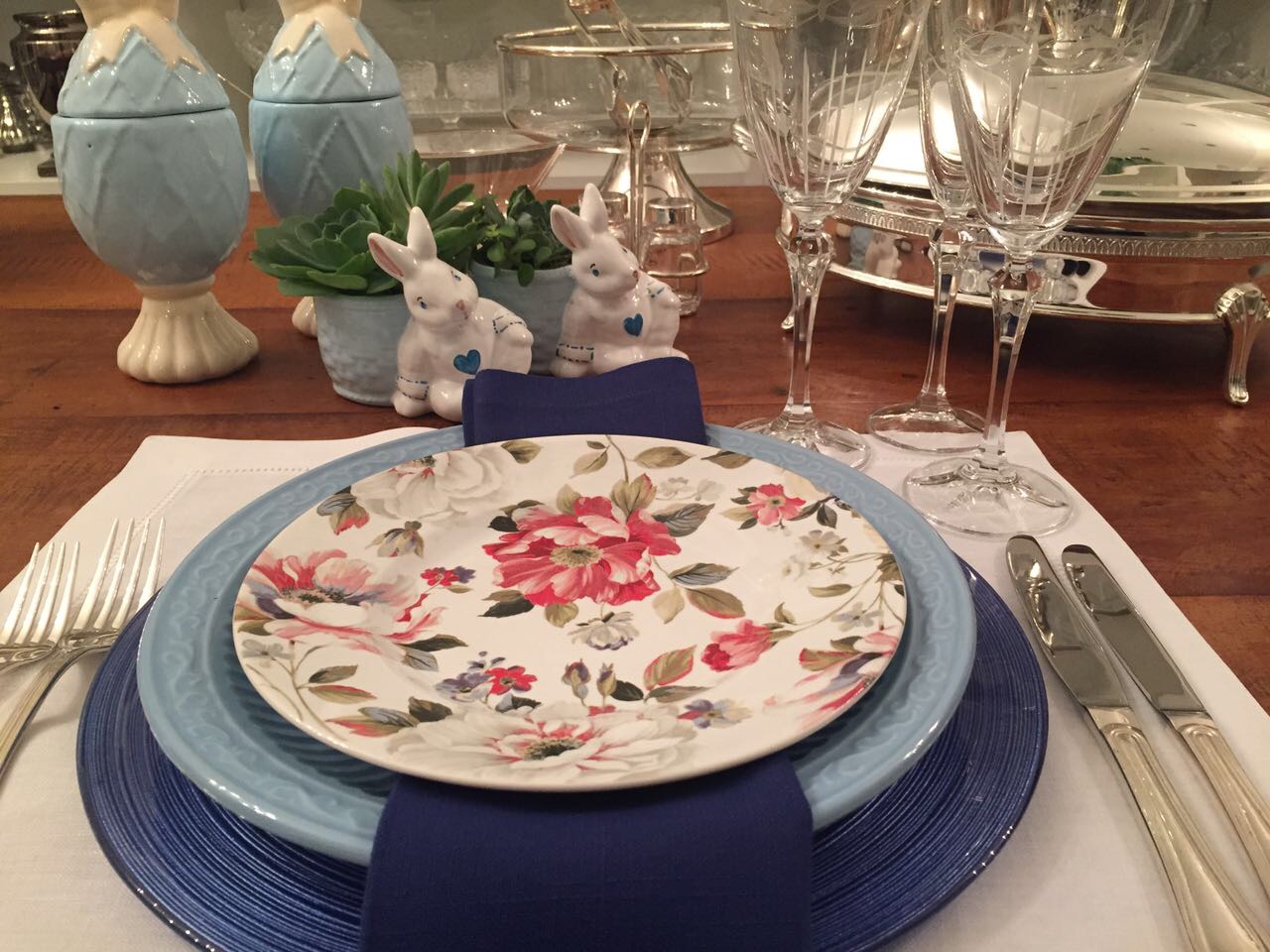 um lugar colocado sobre um sousplat azul claro com pratos rasos de borda azul e sobre ele um prato estampado com flores rosas e vermelhas. Taças de cristal e ovos de porcelana azul ao fundo completam o visual de páscoa.
