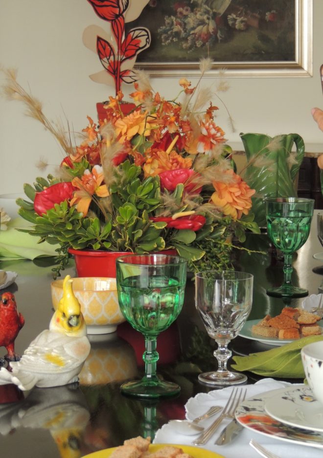 Mesa de jantar montada , com todos os utensilios e acessórios. Flores ao centro na cor branca e laranja. Algumas taças estão em detalhe - a taça de água em tom verde e a taça de vinho branco, em cristal transparente.