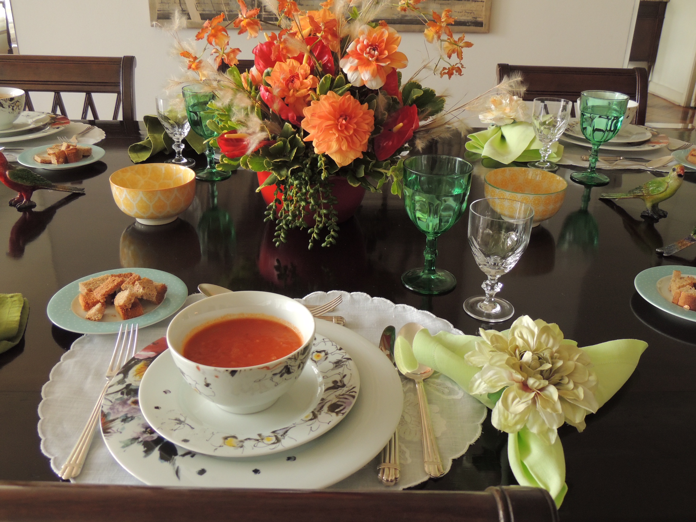 Mesa de jantar montada , com todos os utensilios e acessórios. Flores ao centro na cor branca e laranja. Algumas taças estão em detalhe - a taça de água em tom verde e a taça de vinho branco, em cristal transparente.