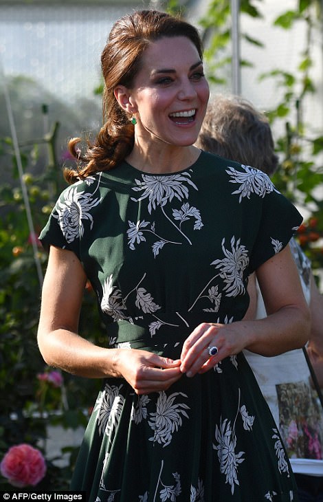 Kate Middleton, em visita a Chelsea Flowers Show, sorri para todos, usando um vestido verde escuro e com detalhes de flores na cor branca, tem seus cabelos presos, Ea está andando , olhando a exposição.