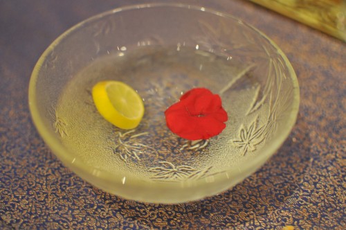 pequena cumbuca de vidro cheia pela metade com água e uma rodela de limão e pétala de flor vermelha para aromatizar.