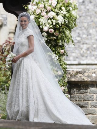 Pippa Middleton vestida de noiva parada junto a porta da igreja antes do seu casamento. Ela segura um pequeno buquê com flores brancas.