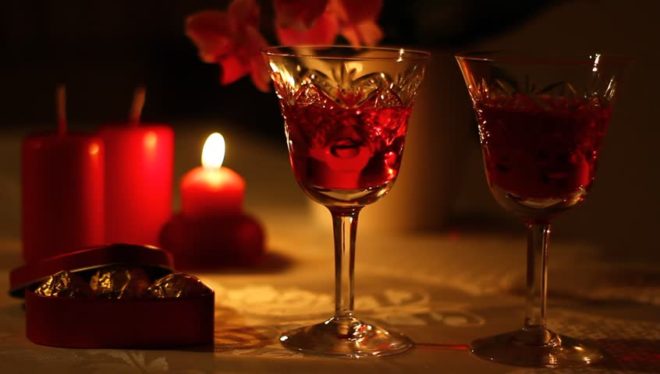 mesa decorada , num ambiente escuro, onde temos em primeiro plano duas taças decoradas, contendo vinho tinto e ao fundo velas decoram a mesa, a menor está acesa.