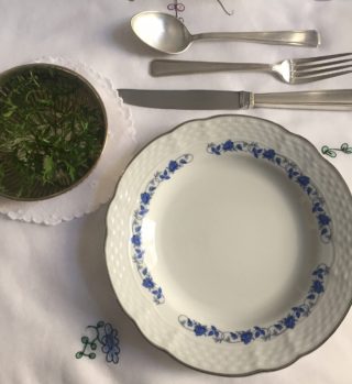 pequena cumbuca de prata com raminhos verdes e água colocada a esquerda do prato de sobremesa e ligeiramente acima