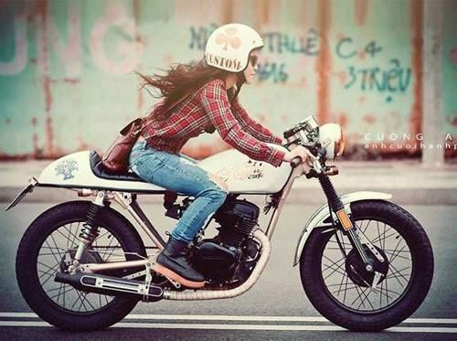 moça sobre uma moto branca com capacete igualmente branco e cabelos castanhos longos esvoaçantes está em alta velocidade. Usa camisa xadrez vermelho, calça jeans e botas pretas de cano curto.