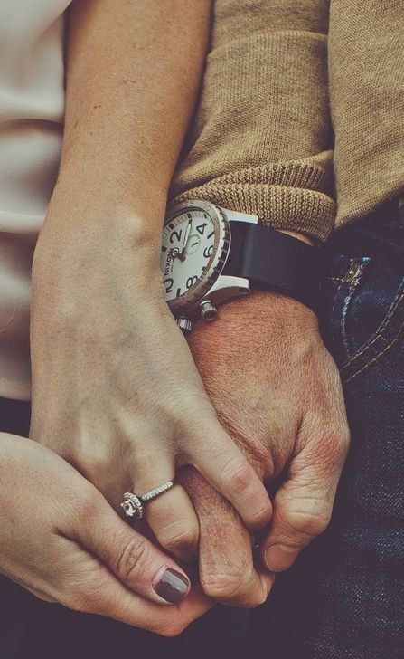 maos de homem e mulher estão entrelaçadas em close na foto. Ela usa um anel no anular e ele um relógio de pulseira de couro preto