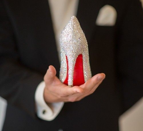 Um homem vestido de terno escuro segura na palma de sua mão um sapato de salto alto todo bordado de cristais - lembrando o sapato da Cinderella nos contos de fadas.