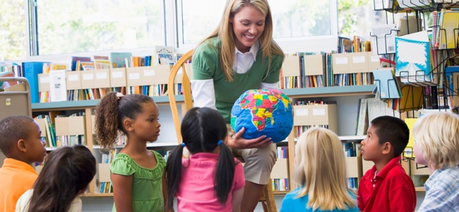 professora bonita, vestida de camisa branca com uma camiseta verde por cima, mostra um globo bem colorido para sete crianças bastante interessadas.Eles estão em uma sala de aula repleta de livros.