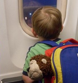 Menino de 3 anos dentro de um avião olhando pela janela. Está com uma mochila colorida nas costas de onde aparece a cabeça de um leão de pelúcia