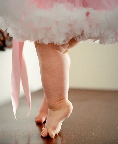 A foto mostra apenas as pernas rechonchudas de uma bebê de 1 ano de idade, usando um vestidinho de festa de tule rosa e branco, descalça sobre o piso de madeira da sala, na ponta dos pezinhos gordos como se estivesse fazendo força para se equilibrar