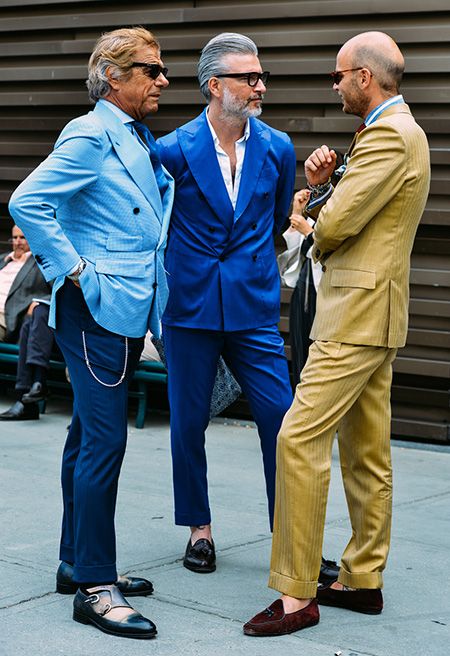 3 homens italianos estão em pé numa praça de Roma, um deles está com blaizer azul claro e calça azul escura , sem meia e sapatos colorido azul e amarelo, o outro com um terno azul petroleo e camisa branca e o outro com terno cor camelo, sapato marrom escuro sem meia.