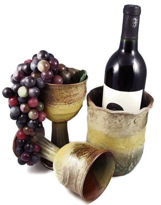 cooler de cerâmica com uma garrafa de vinho dentro, ao lado de duas taças também em cerâmica. uma está em pé com uvas dentro e a outra está caída.