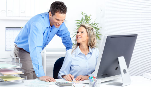 mulher loira bonita, vestida com camisa azul clara, sentada à frente do computador mostra papéis de trabalho para um homem. Percebemos que eles estão em um escritório e tendo uma conversa agradável