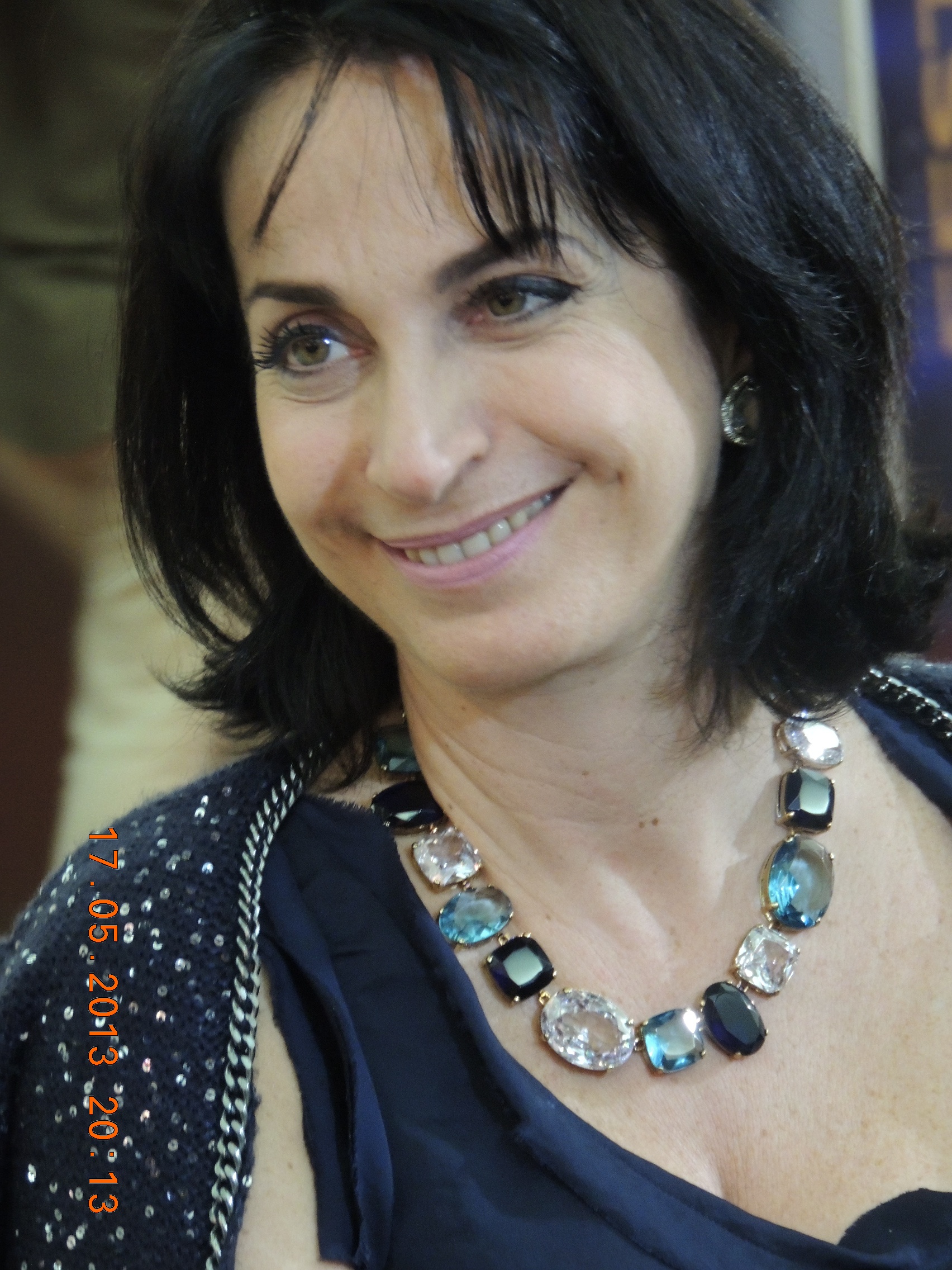 Claudia Matarazzo, seus rosto está inclinado para a direita, sorri, um olhar para o seu interlocutor, usa um lindo brinco colorido e prata junto a orelha, um belíssimo colar de pedra grandes, em varios tons de verde e azul. sua blusa presta de alça, estilo camiseta, por cima veste um lindo casaco com azul escuro com pequenos detalhes azul claro.