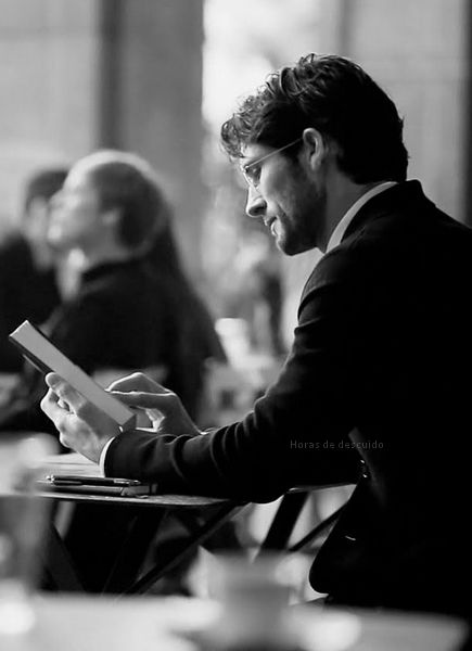 imagem em preto e branco, homem sentado numa mesa de bar, lendo um livro , ele jovem e usa barba