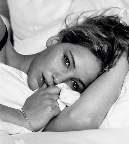 A atriz Jennifer Lawrence, em foto preta e branca, deitada numa cama, seu rosto apoiado no braço esquerdo e o outro segura lençóis, cabelos despenteados,